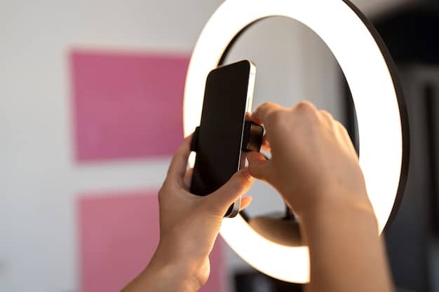 A person holding up a smartphone facing a ring light in a dimly lit room, showcasing how to arrange the lighting, which conveys the user's focus to create appealing visuals. The setting is a bedroom converted into a makeshift studio with soft ambient light, contributing to a cool and modern aesthetic.