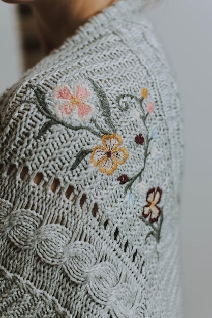 A close-up shot of a woman wearing a sheer, long-sleeved top with delicate floral embroidery. The top is layered over a black lace bralette, and she is wearing high-waisted denim jeans. The background is a blurred urban setting, highlighting the juxtaposition of edgy and feminine elements.
